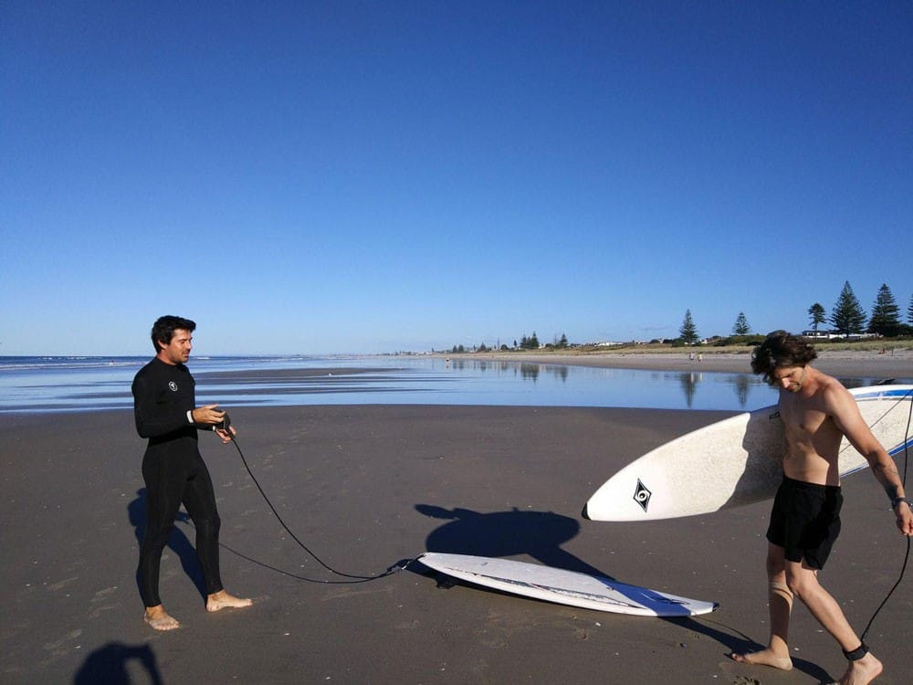 surf en Nueva Zelanda dos chicos con el neopreno en la arena de la playa y dos tablas de surf y de fondo el mar